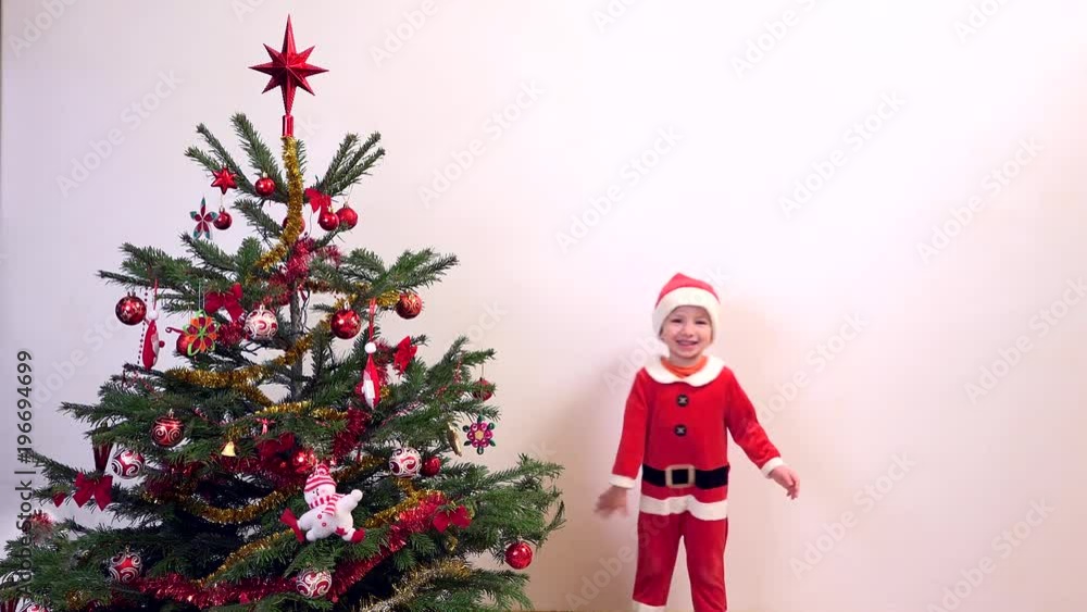 Happy child dressed in Santa Claus jumping near Christmas tree, waiting presents Happy child dressed in Santa Claus jumping near Christmas tree, waiting presents