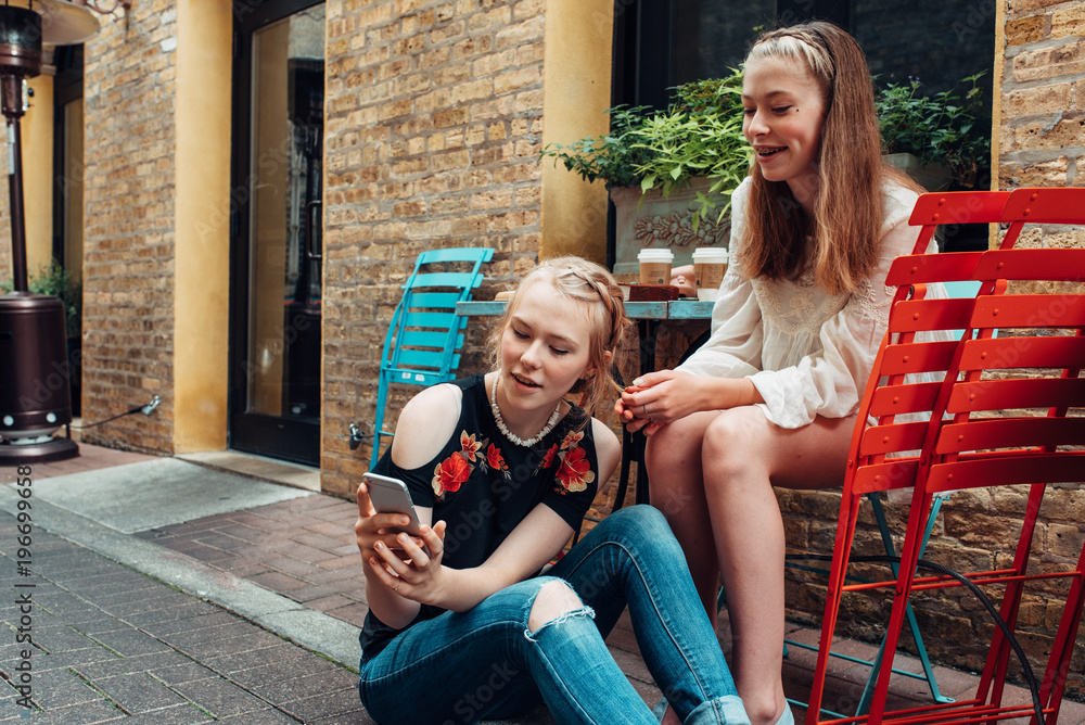 Two young girls checking a smartphone by a cafe table outdoors Two young girls checking a smartphone by a cafe table outdoors