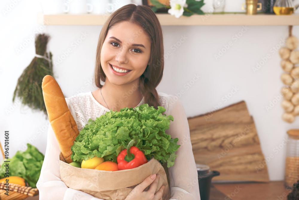Young happy woman holding paper bag full of vegetables and fruits while smiling. Girl have made shopping and ready for cooking in kitchen Young happy woman holding paper bag full of vegetables and fruits while smiling. Girl have made shopping and ready for cooking in kitchen