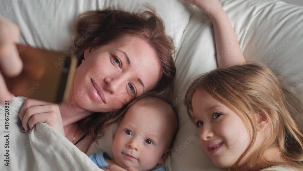 Happy mother and her young children taking selfie with smartphone on bed Happy mother and her young children taking selfie with smartphone on bed