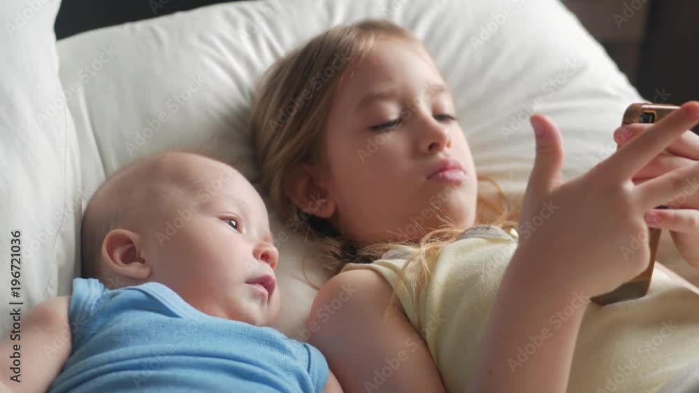 Little cute sister and baby brother is looking or playing with a smartphone on bed at home Little cute sister and baby brother is looking or playing with a smartphone on bed at home