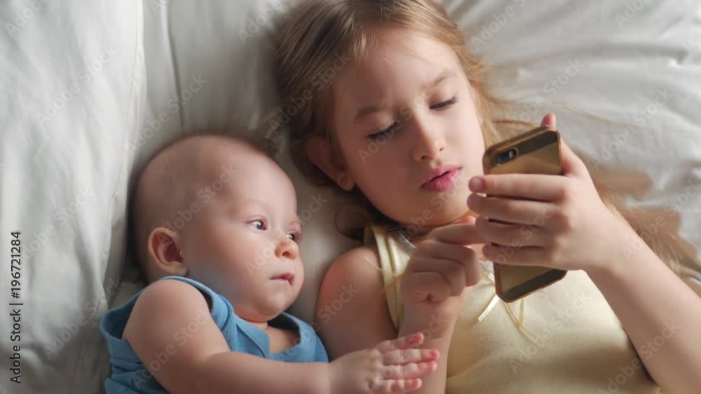 Little cute sister and baby brother is looking or playing with a smartphone on bed at home Little cute sister and baby brother is looking or playing with a smartphone on bed at home