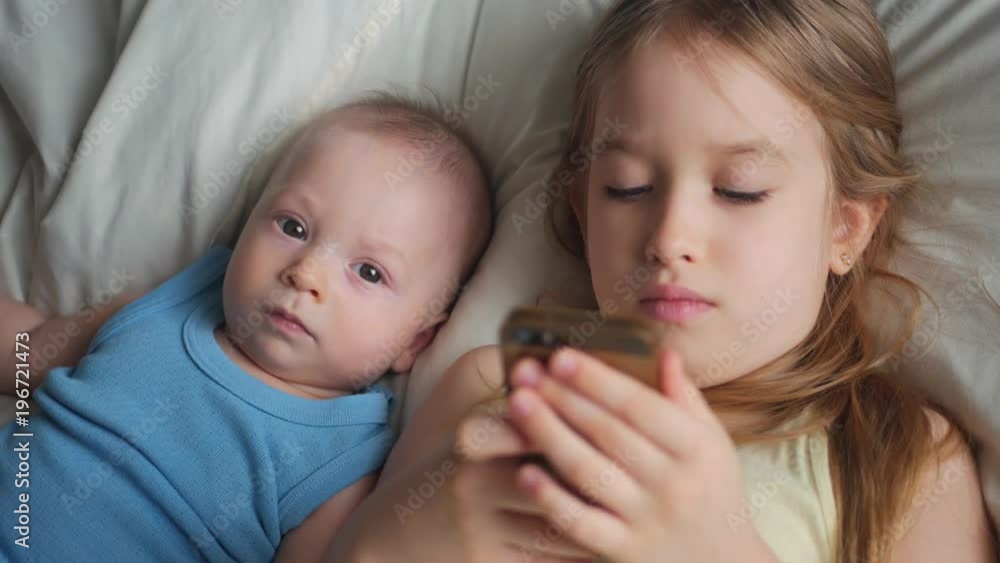 Little cute sister and baby brother is looking or playing with a smartphone on bed at home Little cute sister and baby brother is looking or playing with a smartphone on bed at home