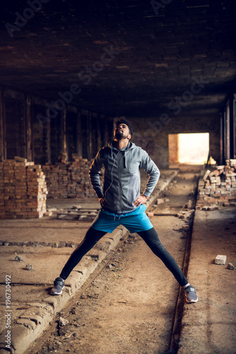 Portrait of active motivated afro-american young attractive athletic man with earphones standing and doing stretching workout with legs open inside of the abandoned place. photo