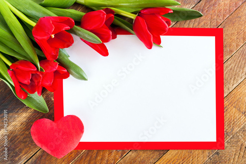 Fototapeta Naklejka Na Ścianę i Meble -  White blank sheet in red frame, red tulips and plush heart on rustic wooden background.