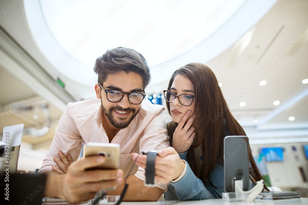 Young cute stylish charming couple testing the new model of gadgets from the desk in the tech store. Young cute stylish charming couple testing the new model of gadgets from the desk in the tech store.