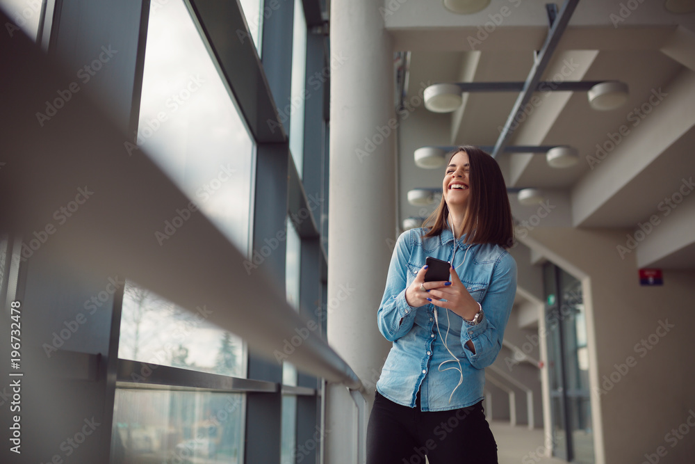 Confident beautiful business woman smiling and using smartphone in light office hall. Smiling girl listening to the music on airport.