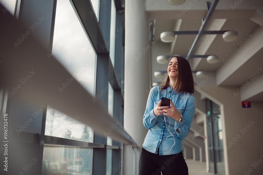 Confident beautiful business woman smiling and using smartphone in light office hall. Smiling girl listening to the music on airport. Confident beautiful business woman smiling and using smartphone in light office hall. Smiling girl listening to the music on airport.