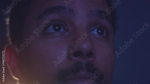 surprised mixed race man watching tv in darkness with different color light from tv on face. Eyes closeup