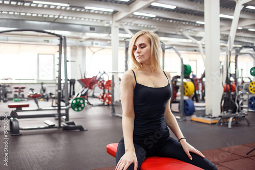 Young slender woman in gym posing against a lot of fitness equipment on background. She wears black T-shirt, pants and sneakers