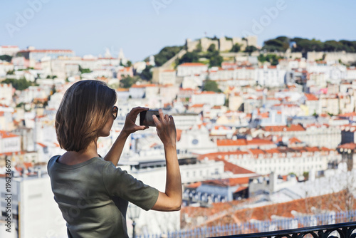 Young beautiful woman holding smartphone and taking picture of Lisbon city, Portugal