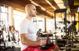 Close up side view of motivated and focused strong muscular active healthy young bald man sitting on the bench with dumbbells in hands and listening music in the sunny modern gym.