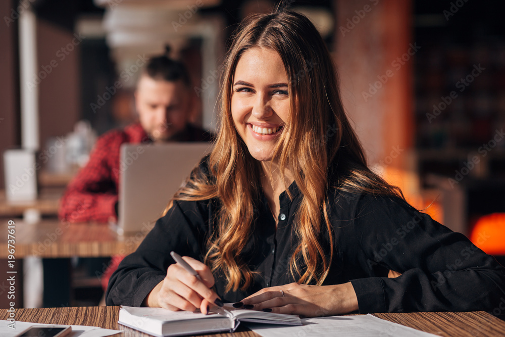 Portrait of a confident woman taking notes in a notepad while sitting at the table in a cafe indoors and looking at camera Portrait of a confident woman taking notes in a notepad while sitting at the table in a cafe indoors and looking at camera