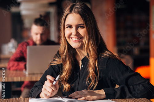 young woman smiling at camera while working remotely in coffee shop. Positive female with notepad