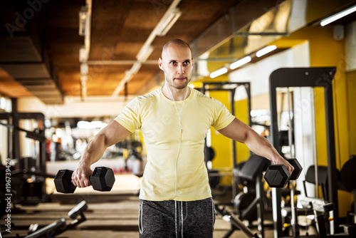 Portrait of strong muscular active healthy young man raising plastic dumbbells with open arms and listening music in the gym.