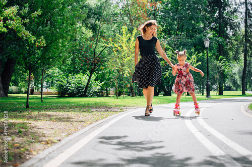 Obraz na płótnie Mother teaching daughter to ride on rollers. Cheerful young woman and cute little daughter ride on roller skates in park. Happy parenting concept. w salonie