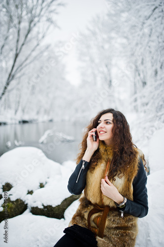 Elegance curly girl in fur coat with mobile phone at snowy forest park at winter.