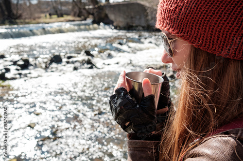 Redhead long haired ginger girl in leather jacket, knitted red cap and sunglasses holding steel cup of hot coffee or tea and smiling near mountain streamy river in spring in sunny weather photo