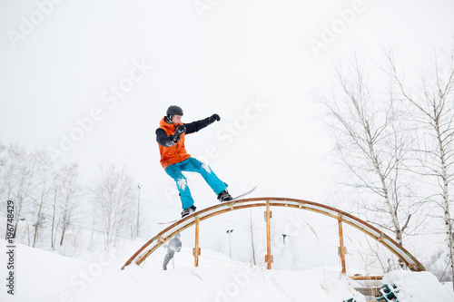 Photo of young sportive man skiing on snowboard with springboard against background of trees