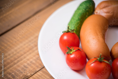 Cherry tomatoes, cucumbers and raw sausages on white plate on wooden table closeup