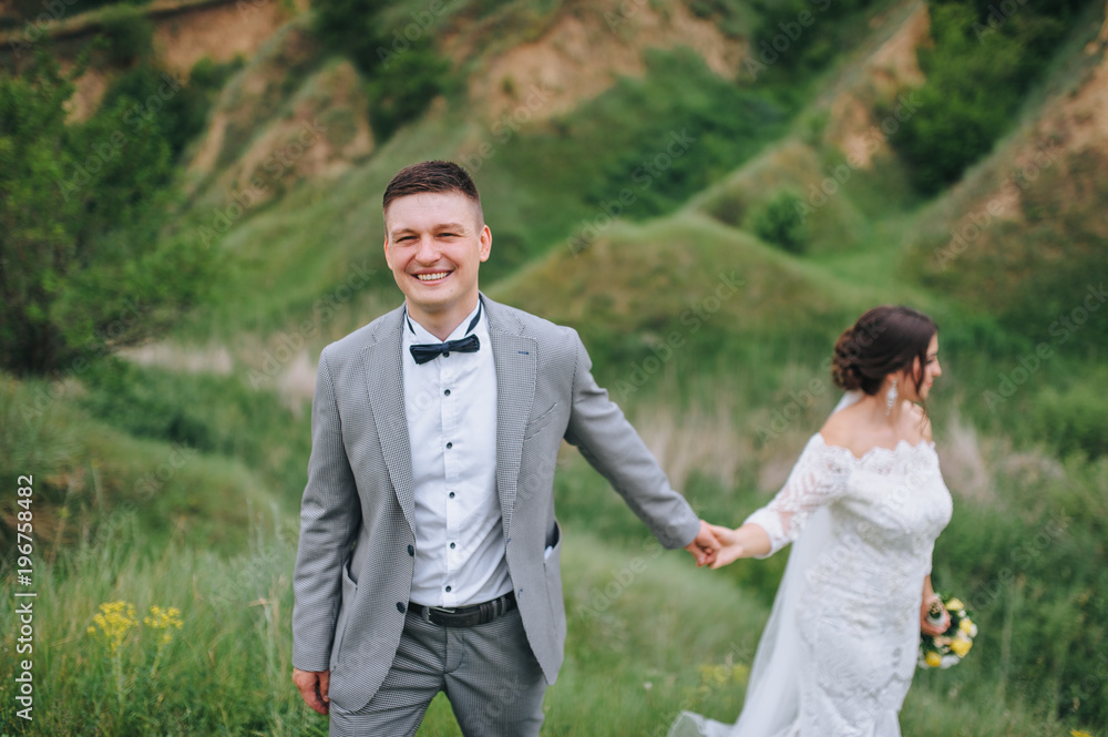 The newlyweds stand against the backdrop of steep hills and a green glade, holding hands. They look in different directions. The newlyweds stand against the backdrop of steep hills and a green glade, holding hands. They look in different directions.