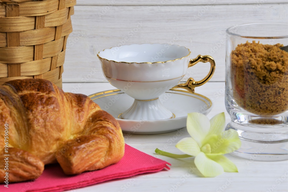 An empty coffee cup on a saucer, Croissant, item basket, flower, brown sugar on a wooden painted background An empty coffee cup on a saucer, Croissant, item basket, flower, brown sugar on a wooden painted background