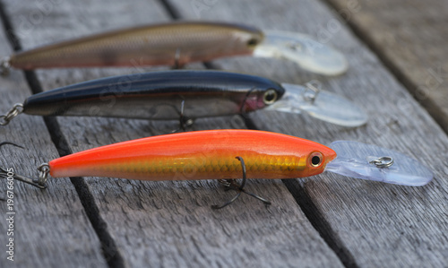 The colorful fish baits are on a wooden background. They are a set of artificial tackles with sharp hooks. There is a close-up fishing texture with plastic wobblers. It is a lot of the models crankbai