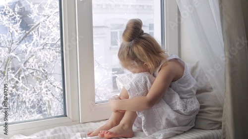 Sad little girl looking through the window sitting on window sill. Winter outside