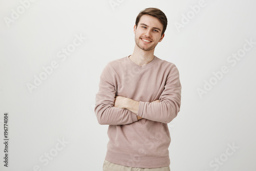 Indoor portrait of positive confident young businessman standing with crossed hands and adorable smile, being satisfied over gray background. I achieved everything by myself and you can do it too © Cookie Studio