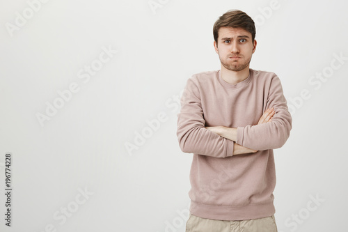 Studio portrait of cute young caucasian male standing with crossed hands in defence pose, sulking and looking with offended expression at camera while standing against gray background.