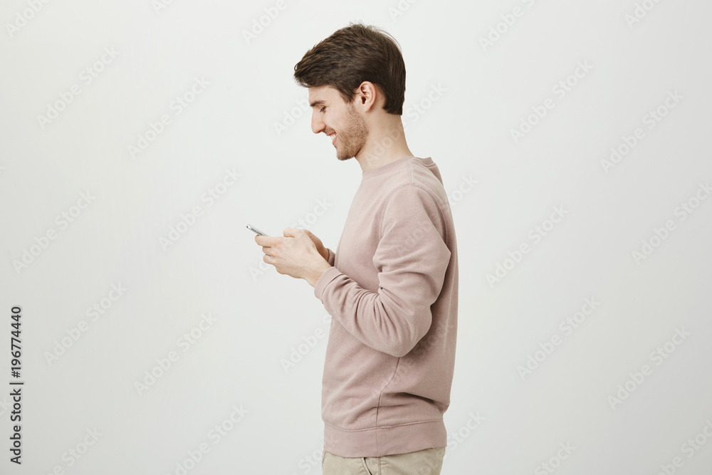 Profile photo of attractive male with trendy haircut and bristle holding smartphone and laughing over something he sees on screen, standing over gray background. Friend send funny photos from party Profile photo of attractive male with trendy haircut and bristle holding smartphone and laughing over something he sees on screen, standing over gray background. Friend send funny photos from party