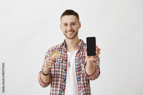 Internet banking and gadget concept. Portrait of handsome happy caucasian with bristle, wearing trendy glasses and showing smartphone at camera while holding credit card and smiling broadly