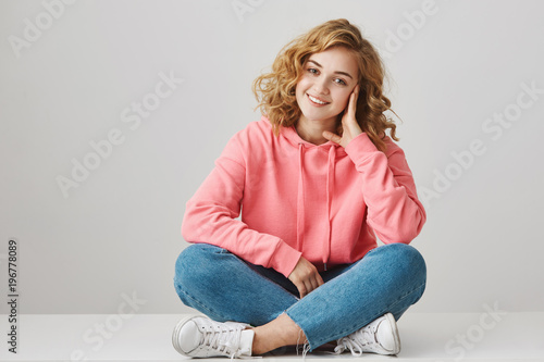 Studio portrait of cute female freelancer with curly hair sitting on floor with crossed lefs leaning head on hand and smiling at camera. Singer with band discuss new song they want to release