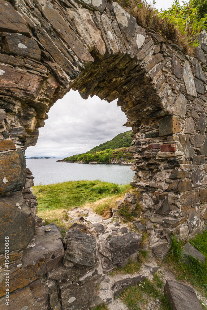 Stromeferry, Scotland - June 10, 2012: Closeup of Window in rock wall of Castle Strome ruins on green hill. Gray blue sky. Green weeds on wall. Gray Loch Carron. Hills seen through the aperture.