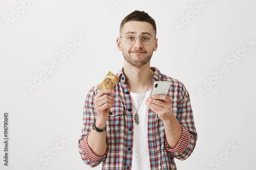 I should buy my girlfriend present. Studio portrait of handsome caucasian guy with beard in glasses, smiling, holding smartphone and credit card while being over gray background.
