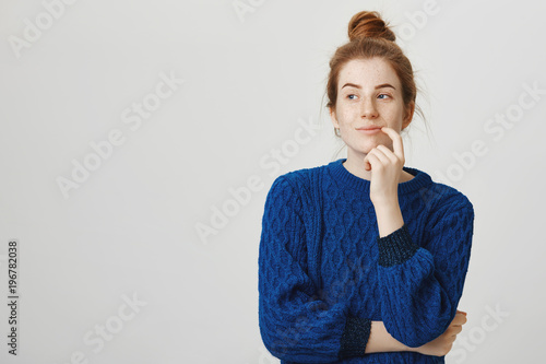 Woman has great plan in mind. Indoor shot of pretty woman with red hair and freckles in winter sweater holding finger on lip and looking aside with slight mysterious smile, thinking or having idea