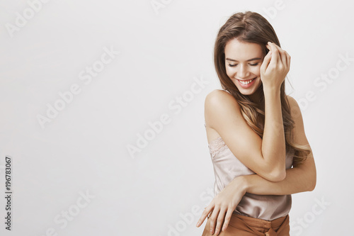 Confident and tender female blushing from compliments. Portrait of flirty sensual feminine woman touching hair near face, looking down and smiling broadly, being sexy and romantic over gray background