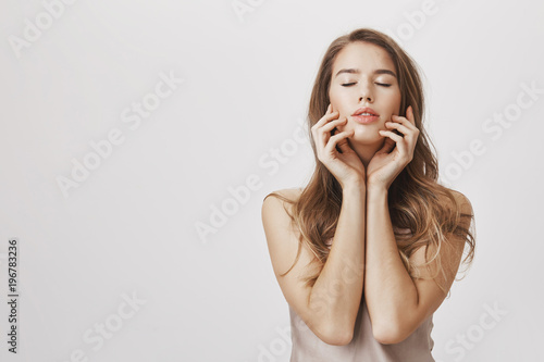 Feeling pleasure and relax after visiting spa. Portrait of dreamy sensual caucasian woman touching face with closed eyes and half-opened mouth, imaging desirable thing, standing over gray wall