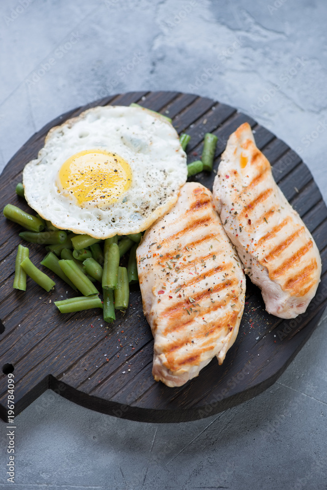 Grilled chicken breast fillet with green beans and fried egg on a black wooden chopping board, selective focus, vertical shot, elevated view