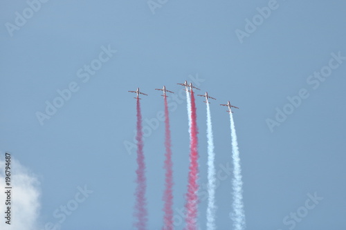Grupa samolotów akrobacyjnych w powietrzu, leci ostro do góry zostawiając za każdym z nich biały i czerwony dym, niebieskie niebo