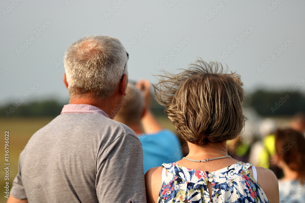 Obraz premium Para osób w dojrzałym wieku, kobieta i mężczyzna, stoją tyłem, w plenerze, patrzą na coś współnie, prezd nimi nieostra postać męzczyzny, wiatr rozwiewa kobiecie krótkie blond włosy