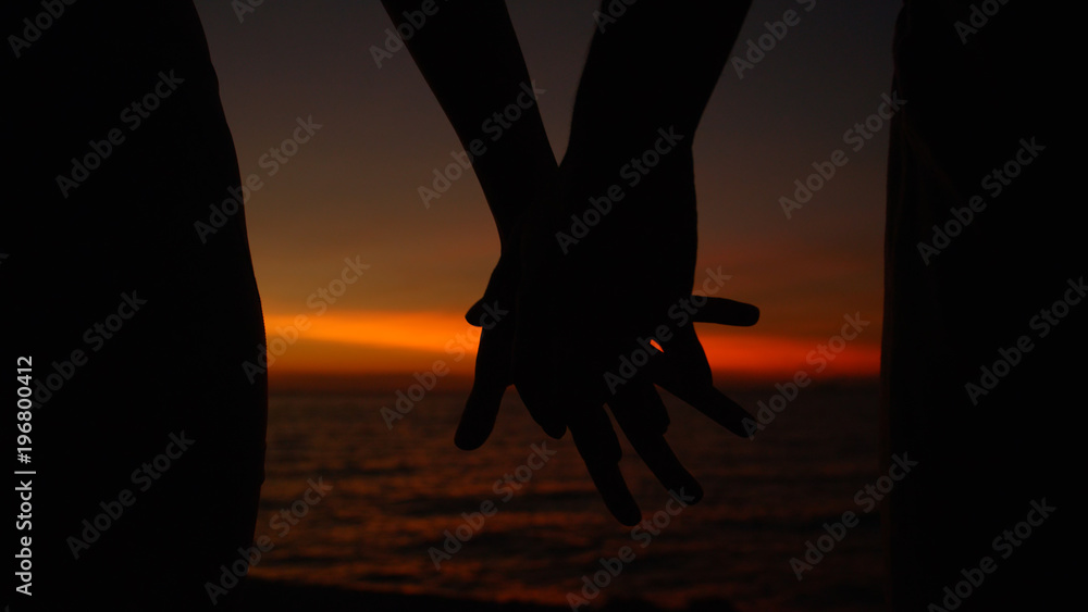 SILHOUETTE: Unrecognizable couple holds hands near picturesque ocean at sunrise.