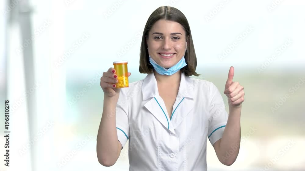 Young female doctor on blurred background. Cheerful woman doctor holding container with pills and gestuing thumb up. Smiling young pharmacist.
