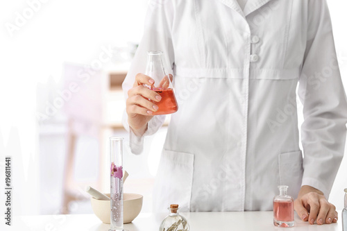 Woman in lab coat holding glass flask with perfume oil near table indoors