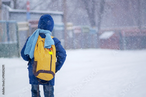 kid (boy) in blue jacket and blue scarf with yellow bagpack going to school in winter nasty blizzard weather, view form behind, copy space