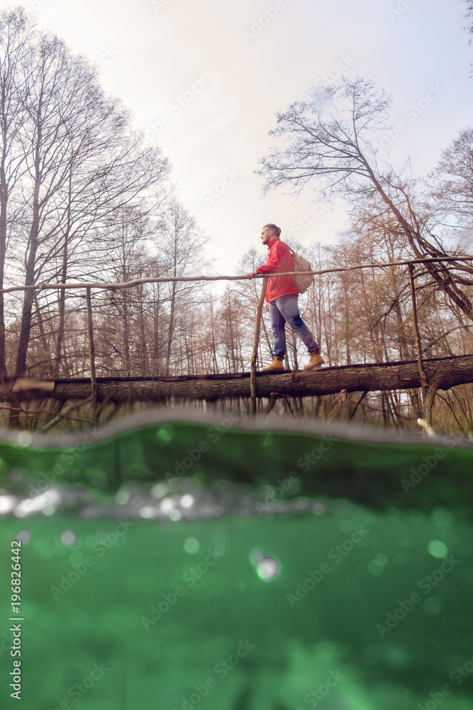 bearded man tourist with backpack in red waterproof jacket crossing forest river over the wooden bridge in spring bearded man tourist with backpack in red waterproof jacket crossing forest river over the wooden bridge in spring