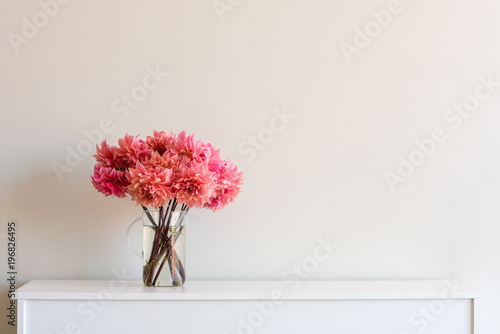 Fototapeta Naklejka Na Ścianę i Meble -  Bright coral pink dahlias in glass jug on white sideboard against neutral wall background