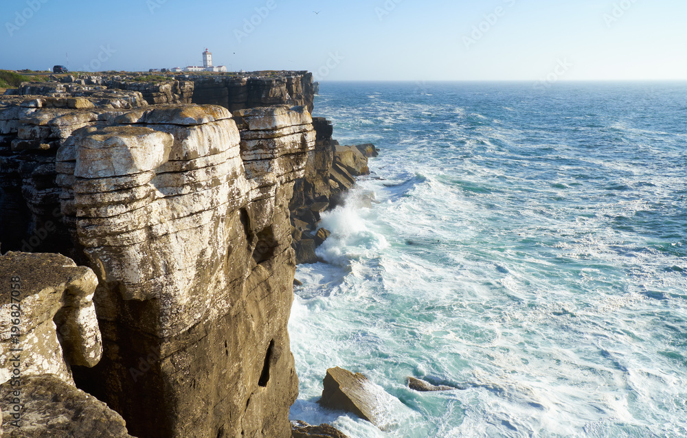 Obraz premium Rocks and waves of surf in the ocean near Cabo Carvoeiro, Peniche peninsula with Lighthouse on background, Portugal