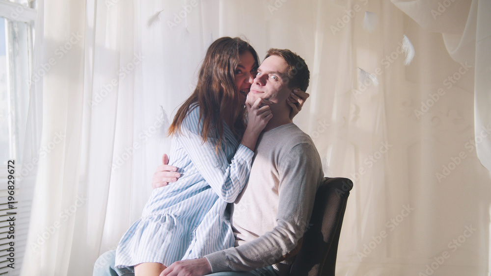 Young guy and girl sitting by the window in a brightly lit room, hugging and kissing