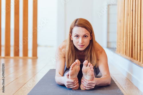Middle aged blond woman practicing yoga, sitting in Seated forward bend exercise, paschimottanasana pose, working out, wearing sportswear, grey pants, indoor, home interior wooden background photo